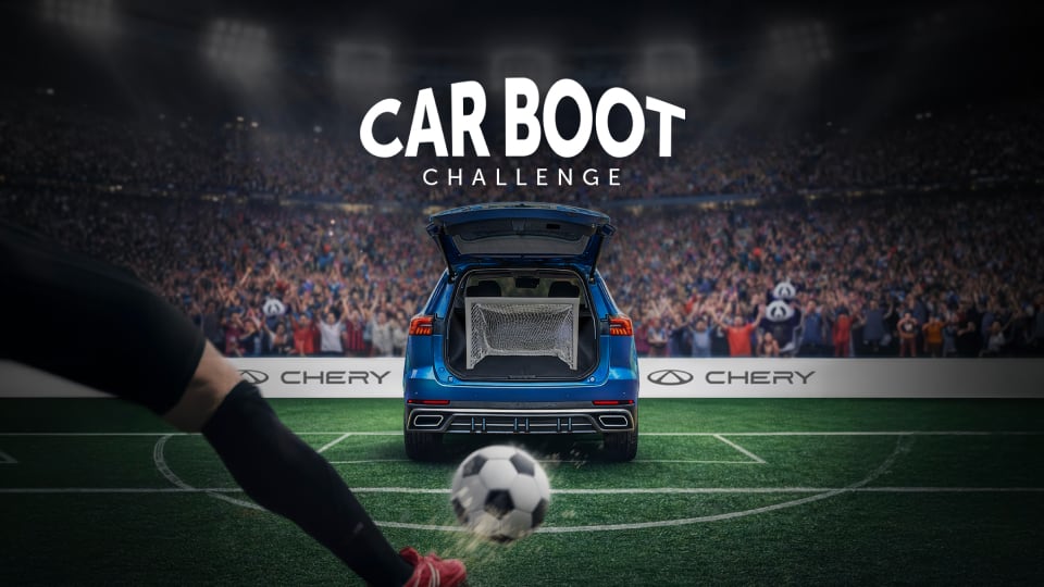 A soccer player kicks a ball towards a blue car with its boot open containing a goal net, in a stadium filled with cheering fans