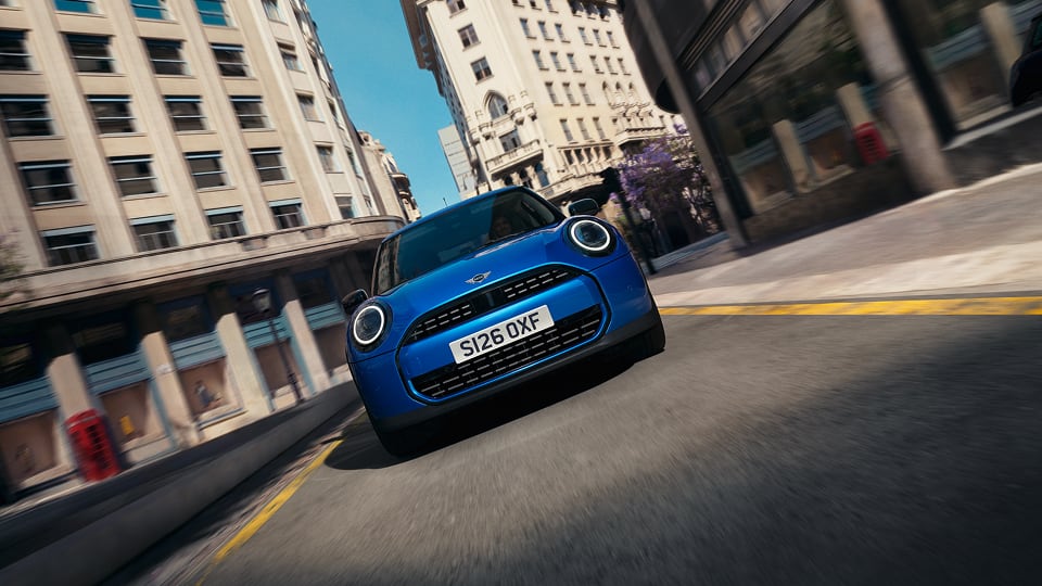 Front view of a blue Mini Cooper driving fast on a city street lined with tall buildings under a clear sky