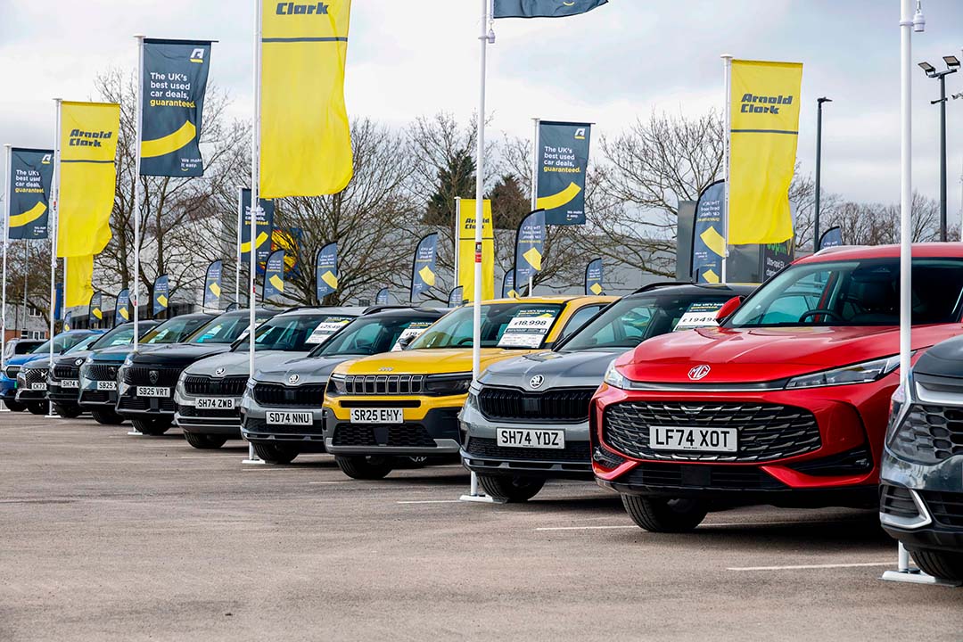 Used cars lined up at a dealership forecourt with promotional flags and price displays.