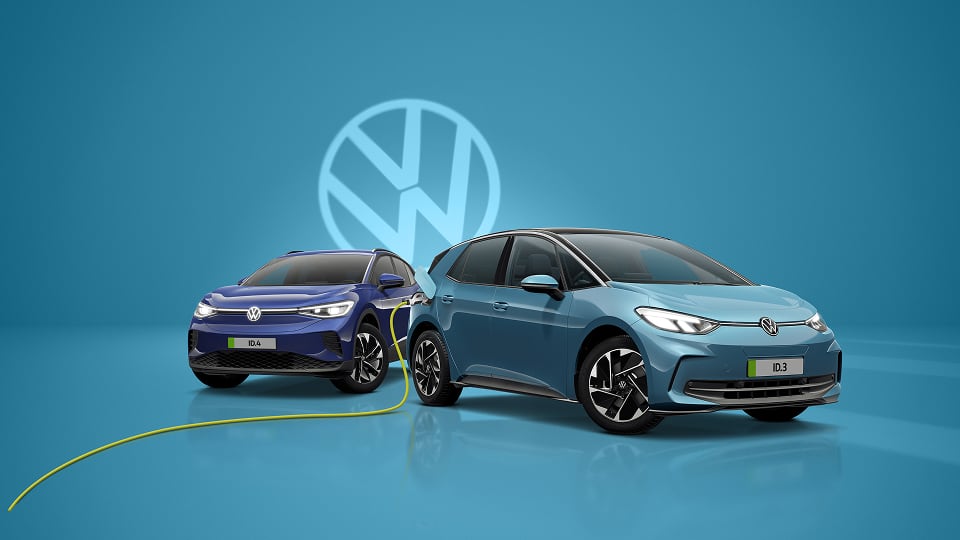 Two Volkswagen electric cars, a blue ID.3 charging and a blue ID.4, with a large VW logo in the background on a blue surface