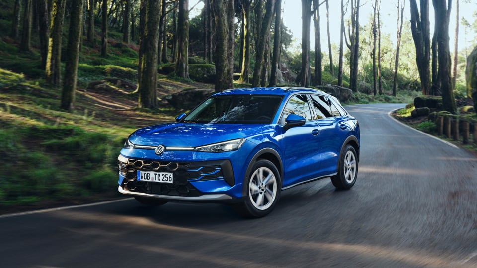 Blue Volkswagen T-Roc driving on a winding forest road surrounded by tall trees and greenery in daylight